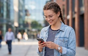 Smiling woman in glasses and a denim jacket reading a text message on her phone while standing on a city street.