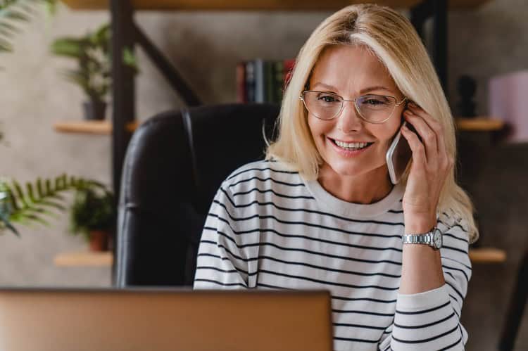 Smiling woman with blonde hair and glasses talking on her phone while looking at her laptop in a home office.