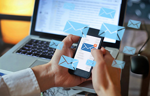 Hands holding a smartphone showing an email icon with seven unread messages, surrounded by floating envelope icons, in front of a laptop.