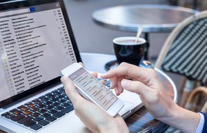 Person checking email on a smartphone at a café table, with a laptop showing a full inbox beside a cup of coffee.