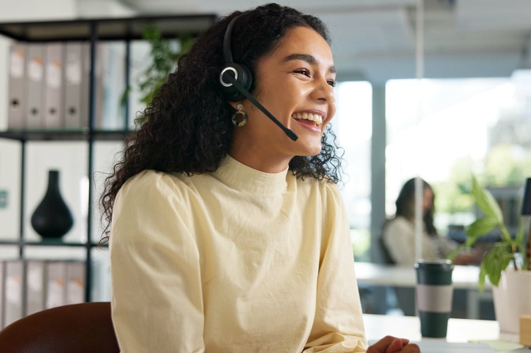 Laughing customer service agent with curly hair wearing a headset at her desk in a bright office.
