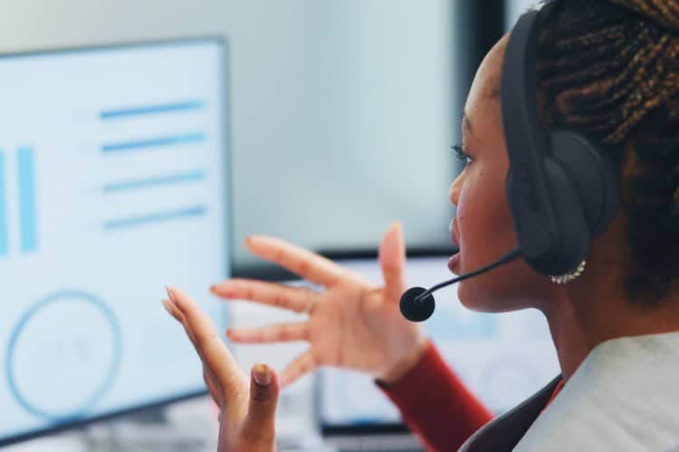 Customer service agent wearing a headset, gesturing while she speaks on a call in front of a monitor showing charts and statistics.